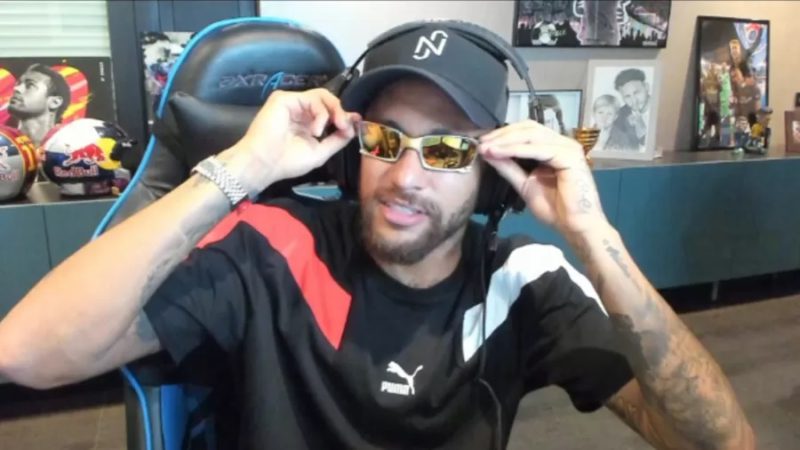 Neymar com um boné, headset e óculos de sol sentado em uma cadeira gamer fazendo pose para a câmera